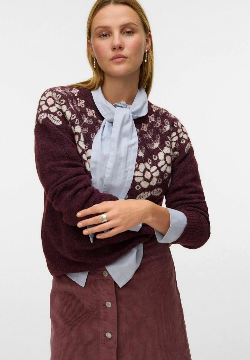 Pull en tricot bordeaux avec un motif floral blanc sur les épaules, superposé à une chemise bleue clair, associé à une jupe en velours côtelé bordeaux.