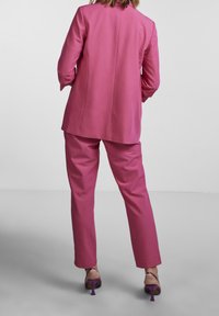 Růžový tailored oblek s dlouhým blazerem a rovnými kalhotami. Hladká textura látky a minimalistický design. Viditelné vysoké podpatky v purpurové barvě.