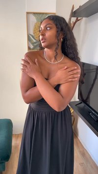 Abito nero senza maniche con corpino aderente e gonna fluida. La modella ha lunghi capelli ricci e indossa una collana di perle, con le braccia incrociate. Pavimento in legno visibile.