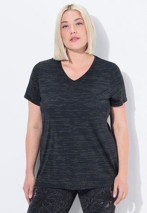 Blonde vrouw met een bobkapsel die een zwart T-shirt met korte mouwen en een V-hals draagt, voorzien van subtiele horizontale strepen, en donkere leggings met een bloemenpatroon.