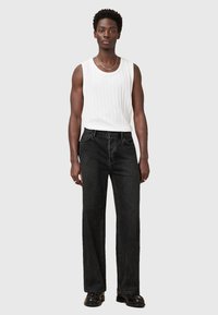 Weißes geripptes Tanktop, schwarze weit geschnittene Jeans, schwarze Stiefel. Silberne Kettenhalskette. Model steht vor einem neutralen Hintergrund.