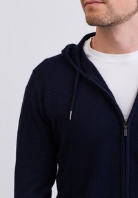 Marinblå zip-up hoodie i texturerat material med huva, dragsko och framfickor. Bärs över en vit T-shirt.