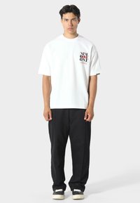 Mladý muž s krátkými tmavými vlasy, oblečený v bílém tričku s grafikou, volných černých kalhotách a černo-bílých teniskách, stojící proti jednobarevnému pozadí.