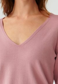 Primo piano di una persona che indossa un maglione a V color malva, mostrando il collo, la parte superiore del petto e parte delle spalle.