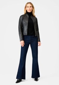 Schwarze Lederjacke mit taillierter Passform, über einem schwarzen Rollkragenpullover getragen. Kombiniert mit dunkelblauen Schlagjeans und schwarzen Stiefeletten mit Absatz.