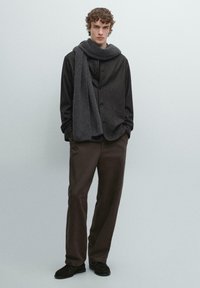 Ciemnoszara kamizelka z mieszanki wełny na czarnej koszuli z długim rękawem, w połączeniu z brązowymi spodniami i prążkowanym szarym szalikiem. Czarnie buty dopełniają strój.