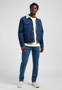 Chaqueta de denim en azul oscuro con un collar forrado de felpa, cierre de botones y dos bolsillos frontales. Combinada con vaqueros de color claro y zapatillas deportivas.
