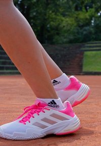 Piernas vistiendo zapatillas deportivas Adidas blancas y rosas y calcetines Adidas blancos en una pista de tenis de arcilla con vegetación de fondo.