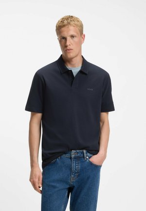 Vaaleahiuksinen mies, jolla on yllään tummansininen lyhythihainen BOSS-logolla varustettu poolopaita ja siniset farkut, seisoo käsi taskussa.