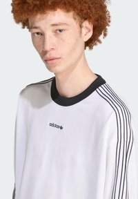 Fehér, hosszú ujjú ing fekete bordás gallérral és mindkét vállon három fekete csíkkal. Fekete Adidas logó a mellkason.