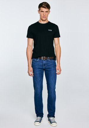 Jauns vīrietis stāv, ģērbies melnā "BIG STAR" T-kreklā, zilās džinsās ar brūnu jostu un zilos sporta apavos ar baltām šņorēm.