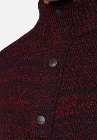 Pull tricoté en rouge foncé et noir, avec un col montant et trois boutons noirs à l'avant, arborant un motif texturé sur toute sa surface.