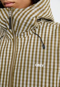 Olivengrønn og hvitstripete jakke med høy krage, som har glidelås, snor-detalj og synlig "OBEY"-logo.