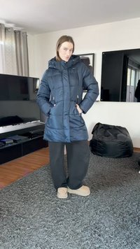 Casaco acolchoado azul-marinho com capuz, bolsos com fecho éclair e textura acolchoada. Combinado com calças pretas largas e calçado bege.