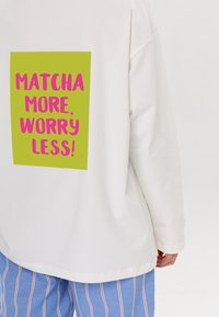 Weißes Langarmhemd mit einem großen grünen Rechteck und pinkem Text, der "MATCHA MORE, WORRY LESS!" liest. Die Rückansicht zeigt eine entspannte Passform.