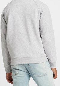 Felpa grigia in cotone con colletto e polsini a costine. Presenta maniche raglan e un orlo dritto. Abbinata a jeans blu chiaro.