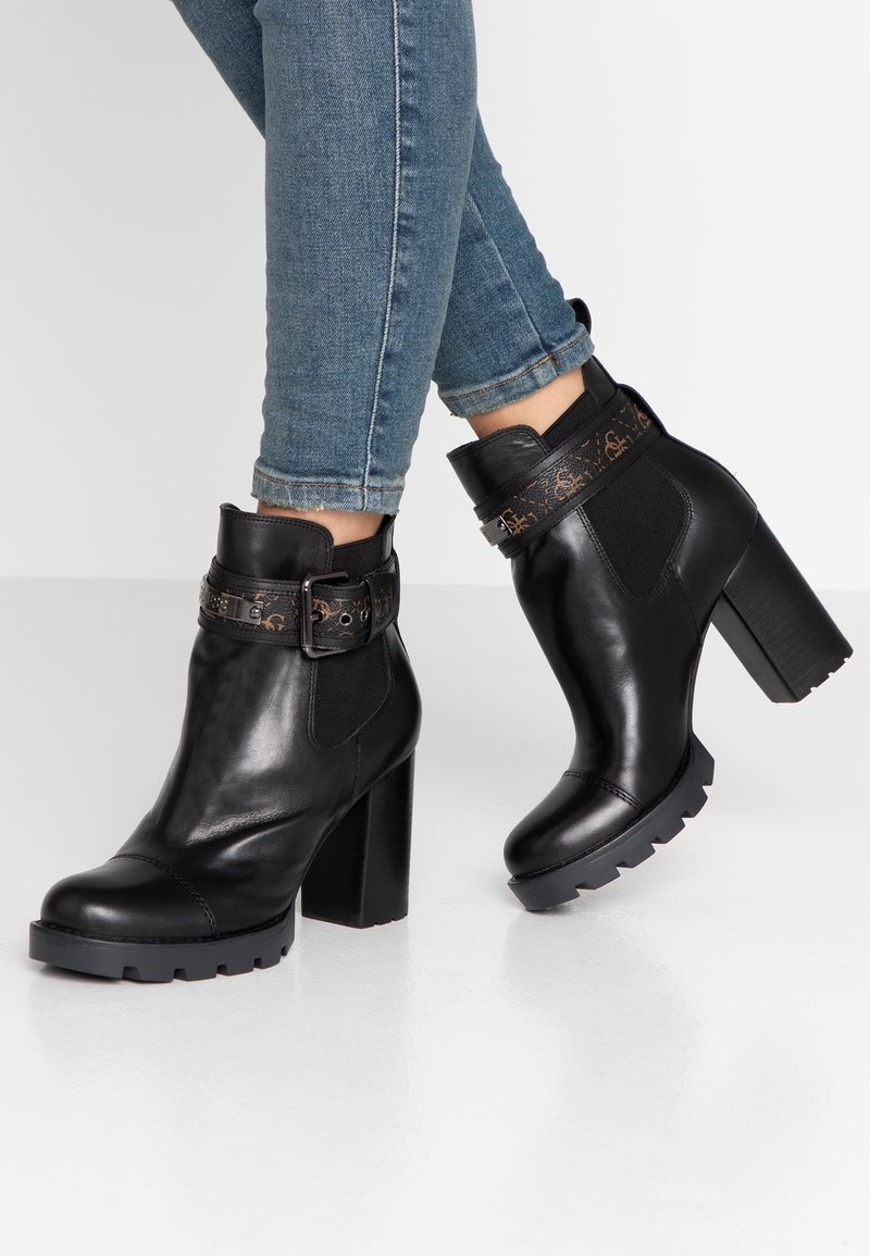 Bottes basses en cuir noir avec talons carrés et boucles décoratives portées avec un jean bleu retroussé sur fond blanc.
