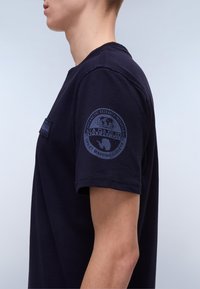 Navy blue cotton t-shirt with a crew neckline, featuring a textured patch on the chest and a logo print on the left sleeve.