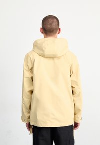 Giacca impermeabile gialla con cappuccio, realizzata in tessuto liscio, dotata di una vestibilità rilassata e polsini elastici, indossata sopra pantaloni scuri.