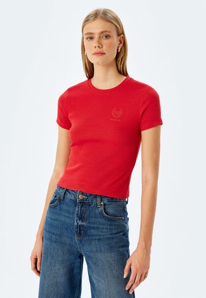 Rød ribbet kortærmet T-shirt med en tætsiddende pasform og rund halsudskæring, med et lille logo i midten. Sættes sammen med blå højhævede jeans.