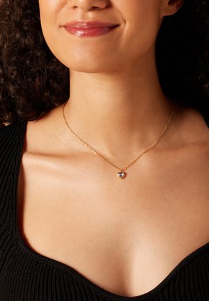 Collier en chaîne dorée avec un pendentif en forme de cœur orné d'un gemme violet. La chaîne est délicate et le pendentif est mis en valeur.