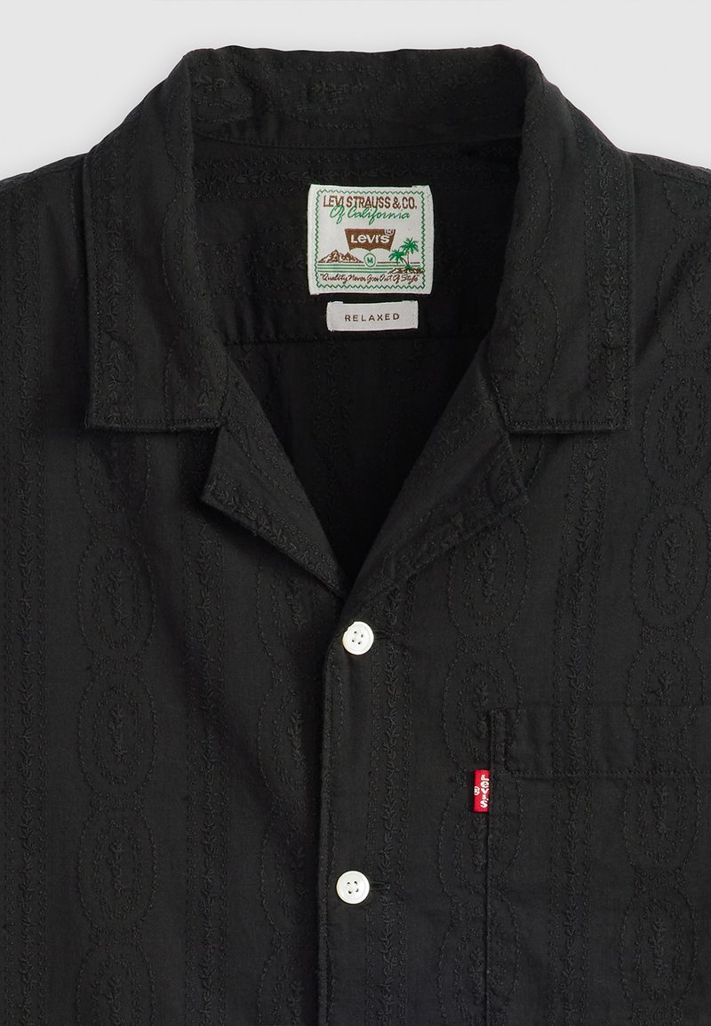 Camisa negra de manga corta con patrones ovalados bordados, dos botones blancos y un bolsillo en el pecho izquierdo que presenta una etiqueta roja de Levi's.