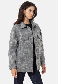Graue Jacke mit strukturiertem Muster, ausgestattet mit einem Kragen, vorderen Taschen und Knopfverschluss. Getragen über einem schwarzen Oberteil und kombiniert mit Jeans.