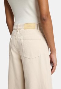 Beige wide-leg jeans lavet af bomuld med et lædermærke, der står "FOR ALL MANKIND," med to baglommer og synlige syninger.