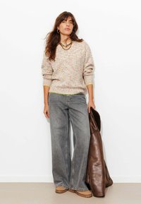 Pull en tricot multicolore avec poignets côtelés, jean large gris et veste en cuir marron. Pose debout, avec des accessoires simples en or.