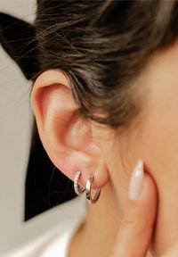 Srebrne kolczyki-hoop wyeksponowane na uchu, jeden z przezroczystymi cyrkoniami. Gładkie, metaliczne wykończenie, podkreślające minimalistyczny design i refleksyjną teksturę.