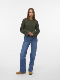 Groene gebreide trui met brede mouwen, gecombineerd met blauwe denim jeans. Bruine schoenen maken de outfit compleet. Schoon achtergrond voor duidelijkheid.