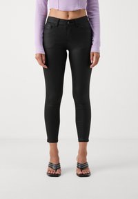 Zwarte skinny jeans van een gladde, rekbare stof. Hoog in de taille met voorzakken, gecombineerd met zwarte sleehakken.