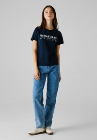 T-shirt in cotone blu navy con il logo "RedBull RACING" in bianco. Abbinato a un paio di jeans in denim blu chiaro e sneaker beige.