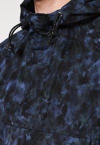 Veste anorak avec un motif abstrait bleu foncé et noir, dotée d'une poche avant, d'une capuche à cordon de serrage et d'une texture de tissu durable.