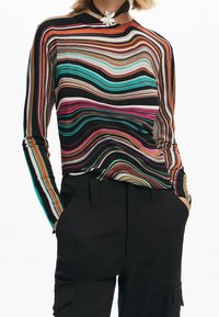 Femme portant un haut à manches longues à rayures ondulées multicolores, un pantalon noir avec poches latérales, et un collier ras du cou avec un grand pendentif en forme de fleur blanche.