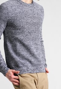 Sweter dziany w odcieniach szarości z teksturowanym wzorem w zygzak, z okrągłym dekoltem i prążkowanymi mankietami, zestawiony z jasnymi spodniami.