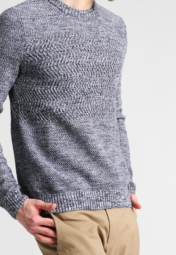 Sweter dziany w odcieniach szarości z teksturowanym wzorem w zygzak, z okrągłym dekoltem i prążkowanymi mankietami, zestawiony z jasnymi spodniami.