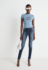 T-shirt aderente azzurro chiaro con grafica di un delfino, abbinato a jeans skinny scuri, tacchi a spillo argentati e una piccola borsa metallica.
