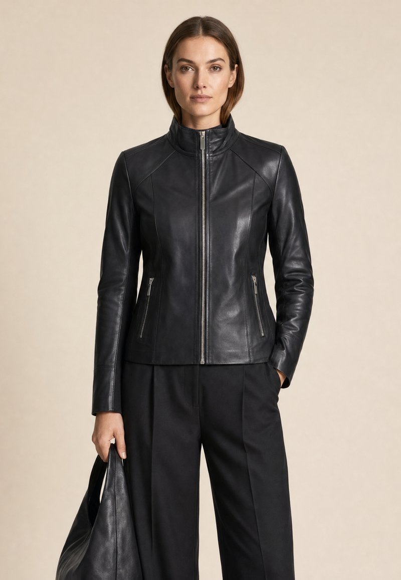 Frau trägt eine eng anliegende schwarze Lederjacke mit Frontreißverschluss, schwarze Hose, hält eine schwarze Lederhandtasche vor einem einfarbigen beigen Hintergrund.