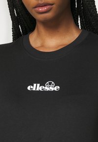 Gros plan d'une personne aux cheveux tressés portant un T-shirt noir avec le logo blanc « ellesse » centré sur la poitrine.