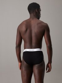 Crne gaćice od mekog materijala s širokim bijelim pojasom na kojem je ispisan logo "Calvin Klein". Model prikazan straga, s kratkom kosom.