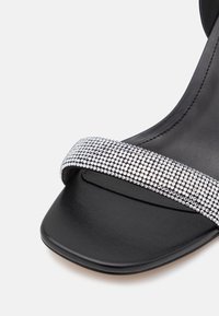 Talons noirs avec un bout carré, dotés d'une bride en strass argenté scintillant. La partie supérieure a une texture lisse, contrastant avec la semelle mate.