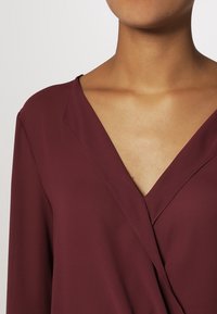 Blusa envolvente burdeos con un escote en V profundo, textura suave y mangas largas. Presenta un sutil caído en la parte delantera.