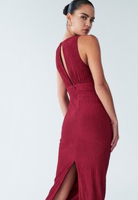 Rdeča obleka z teksturo, oprijetega kroja, z odprtino na hrbtu in stranskim razporkom za lažje gibanje.