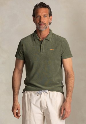 Olijfgroene polo met korte mouwen, kraag met twee knopen en een oranje accent op de borst. gedragen met textuurwitte broek met aantrekkoord.