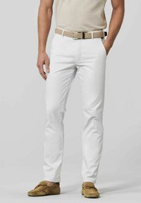Chinos blancs en coton avec une coupe ajustée, arborant une ceinture tissée beige et des mocassins marron en cuir souple. La tenue est complétée par un haut beige clair.