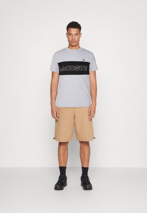 Camiseta de algodón gris con una banda horizontal negra que lleva "LACOSTE" en letras blancas. Combinada con pantalones cortos caqui y zapatos negros.