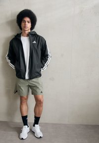 Sort letvægtsjakke med hvide striber, hvid t-shirt, olivengrønne shorts, sorte sokker og hvide sportsko. Enkel design med logoaccentuatior.