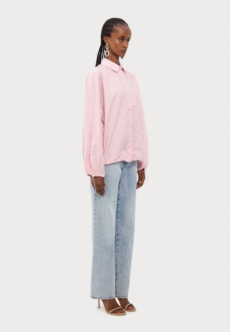 Camisa oversized riscada em rosa e branco, com colarinho e bainha ajustável, combinada com jeans amplos de perna larga em azul claro e sandálias bege.