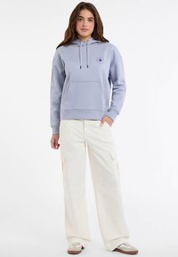 Gaiši zila sporta džemperis ar kapuci, priekšpusē kabata un uzdrukāts zvaigznes grafisks attēls. Kopā ar platām krēmkrāsas biksēm ar kabatām un sporta apaviem.